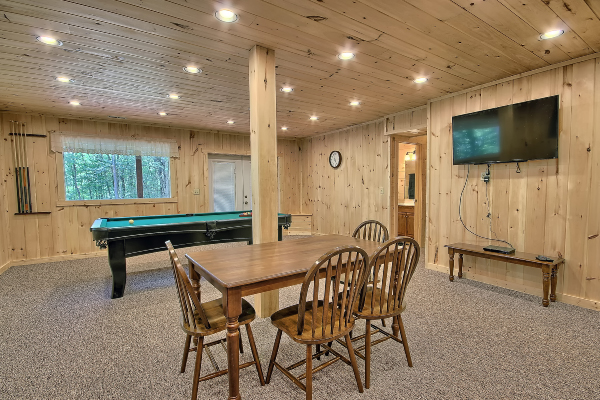 South side of Game Room, in Lower Level, looking North, showing the Table and Chairs, with the TV Screen on right and Pool Table and Pool Cues ahead. Also, Picture Window and Lower Level Walkout Door ahead, with hallway to bedrooms and bath on right.